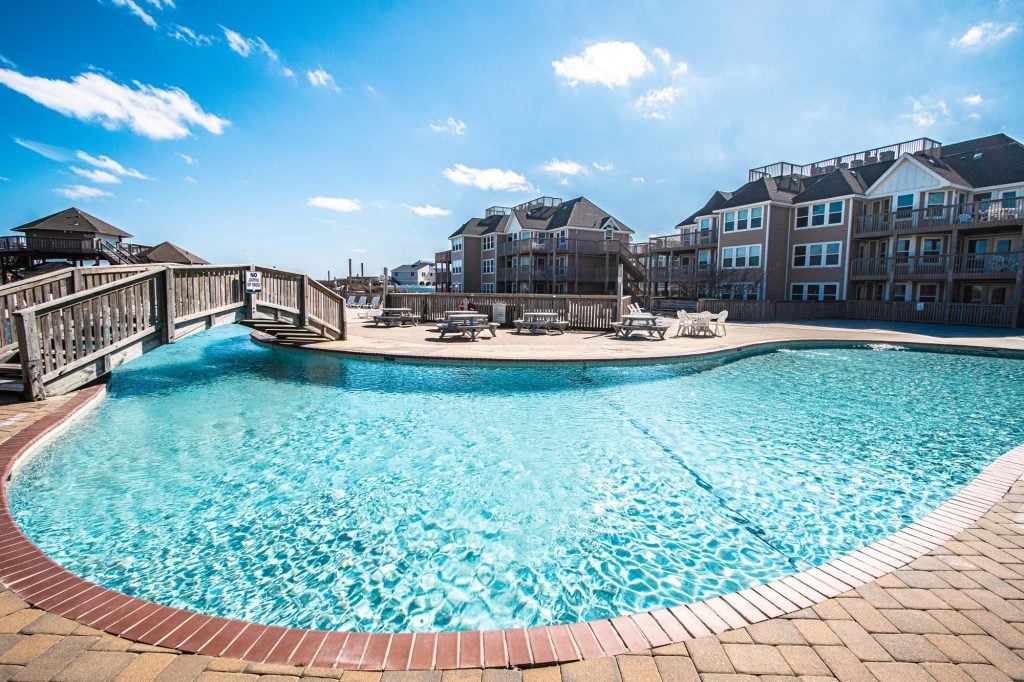 Curved resort pool with turquoise water, brick deck, wooden bridge, and multi-story beach houses under blue sky